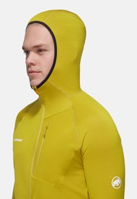 Gul huvjacka med texturerat tyg, ficka med dragkedja på bröstet och logotyp på ärmen. Skräddarsydd passform och integrerad huvddesign.