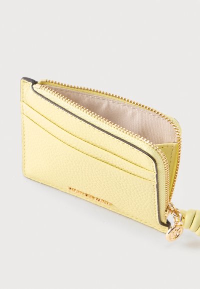 Porte-cartes compact en cuir jaune avec fermeture éclair dorée, fentes pour cartes à l'avant et tire-zip rond doré avec un détail noué.