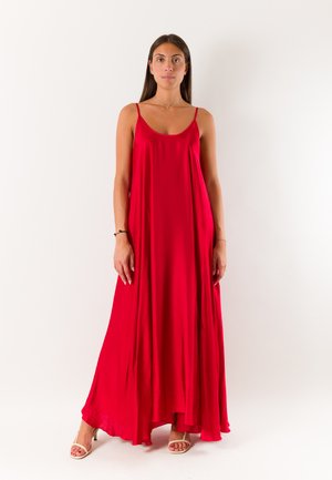 Donna con capelli lunghi e scuri che indossa un vestito rosso fluente senza maniche e tacchi aperti beige, in piedi davanti a uno sfondo bianco semplice.