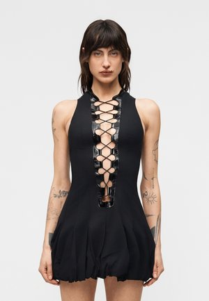 Γυναίκα με σκούρα μαλλιά που φοράει μαύρο αμάνικο φόρεμα με σχέδιο δέσιμου σε σχήμα σταυρού στο μπροστινό μέρος και τατουάζ και στα δύο χέρια, στέκεται μπροστά σε απλό φόντο.