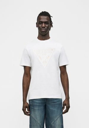 Muž stojí čelom vpred, má na sebe biele tričko s krátkym rukávom s vyblednutým trojuholníkovým logom a modré džínsové nohavice, na jednoduchom pozadí.