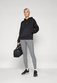 Μαύρο φούτερ με κουκούλα και τσέπη στο μπροστινό μέρος, συνδυασμένο με γκρι κολάν και μαύρα αθλητικά παπούτσια. Κρατάει μια μαύρη τσάντα γυμναστηρίου στο ένα χέρι.