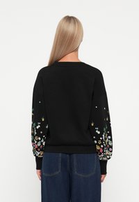 Femme aux longs cheveux blonds portant un sweat-shirt noir avec des motifs floraux multicolores sur les manches et un jean bleu, vue de dos.