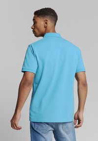 Lichtblauw polo shirt met een geribde kraag, korte mouwen en een rechte zoom, gedragen met lichtblauwe jeans. De stof lijkt glad en casual.