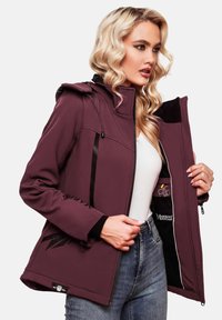 Blonde woman wearing purple softshell jacket, holding it open to show branded interior with black and white details.