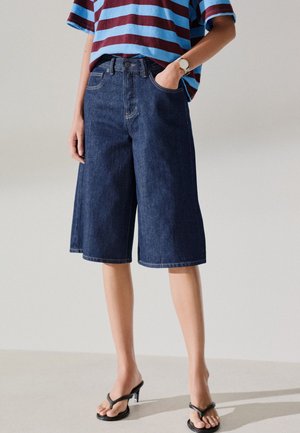Person iført mørkeblå knee-length denim shorts, stribet blå og vinrød skjorte, sorte hælede sandaler og et armbåndsur på venstre håndled.