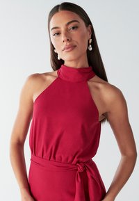 Sarkana halterveida kakla kleita ar augstu apkakli, savelkamu vidukli un sasienamu akcentu. Gluds audums ar pieguļošu dizainu un minimālu rotājumu.