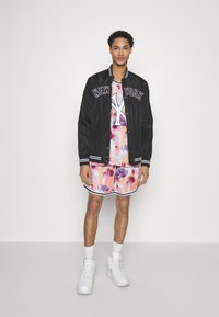 Chaqueta bomber acolchada negra con letras "NEW YORK", camiseta de estampado abstracto multicolor, pantalones cortos rosas con detalles blancos, zapatillas blancas de caña alta.