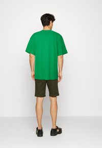 Groene oversized T-shirt met korte mouwen, gecombineerd met donkergroene shorts. Accessoires zijn zwarte sandalen. Glad stofoppervlak.