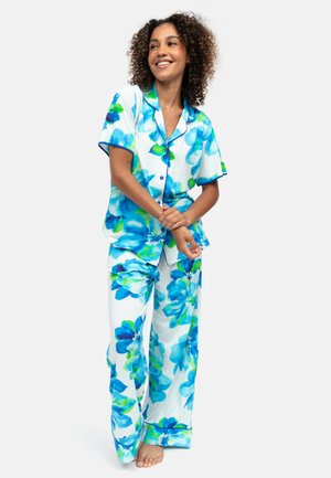 Vrouw die glimlacht en blootsvoets staat, gekleed in witte pyjama met grote blauwe en groene bloemenprints.