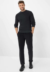 Zwarte gebreide trui met ronde hals en ribbels op de mouwen, gecombineerd met zwarte ribfluweelbroek en zwarte veterschoenen, voor een witte achtergrond.