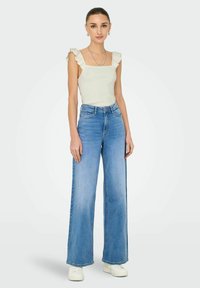 Ribbede, firkantede top i off-white med flæsekanter, parret med højtaljede, vide blå jeans og hvide sneakers.