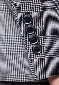 Dopasowana marynarka w kratę w granatowo-białym kolorze. Posiada czarne guziki z wygrawerowanym napisem "Lancerto", ukazując szczegółową teksturę materiału.