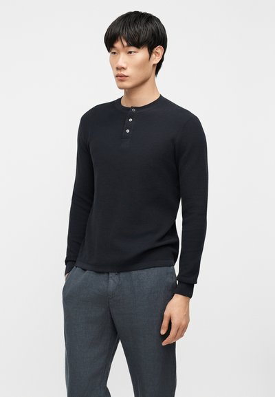 Νεαρός άνδρας που φοράει μαύρο μακρυμάνικο πουκάμισο henley και γκρι παντελόνι, στέκεται με τα χέρια στις τσέπες, κοιτάζοντας προς τα πλάγια.