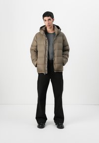 Puffer jacket in muted olive green with horizontal stitching, zip closure, and hood; paired with gray t-shirt and black pants.