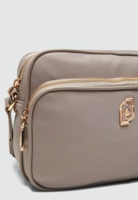 Geantă crossbody bej cu un finisaj texturat, două compartimente cu fermoar și detalii de hardware de ton auriu. Prezintă un accent cu logo pe față.