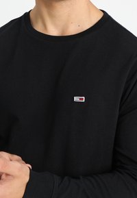 Schwarzes Langarmshirt aus Baumwollstoff mit Rundhalsausschnitt und einem kleinen Logopatch mit rot, weiß und blauen Akzenten auf der Brust.