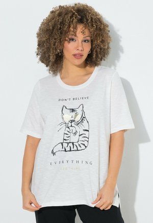 Frau mit lockigem Haar trägt ein weißes T-Shirt mit Tigerzeichnung und Text "Don't believe everything you hear."