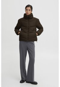 Dunkelbraune Pufferjacke mit hohem Kragen, Reißverschluss und seitlichen Taschen, kombiniert mit grauen, weit geschnittenen Hosen und schwarzen Schuhen.