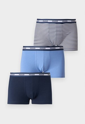 Homme noir athlétique debout pieds nus, portant un boxer bleu clair avec une ceinture de marque, regardant vers sa gauche sur un fond gris uni.