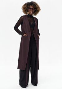 Długa bordowa kamizelka z otwartym przodem i kieszeniami, noszona na przejrzystym czarnym topie i szerokich czarnych spodniach. Przezroczyste okulary przeciwsłoneczne i efektowne kolczyki.