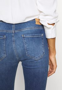 Jeans de mezclilla azul ajustados con bolsillos traseros y un parche de logo de cuero. La tela tiene una textura suave y un ligero desgaste cerca de la costura.