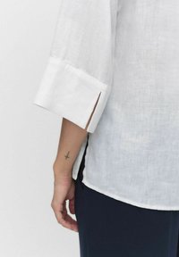 Hvid linnedskjorte med trekvartærmer med slids, parret med navy bukser. Synlig tatovering på håndleddet. Blød tekstur og let udseende.
