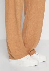 Beige strikkede sweatpants med en afslappet pasform og ribbede manchetter, parret med hvide sneakers med gyldne øjer og en glat overflade.