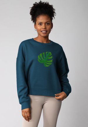 Turquoise sweatshirt met een groene monstera-bladprint, met een relaxte pasvorm en geribbelde manchetten, gecombineerd met licht geribbelde leggings.