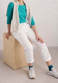 Turkos långärmad skjorta med en ljusgrå tröja över axlarna, vita uppvikta byxor och vita sneakers med turkosa strumpor, sittande på en träblock.