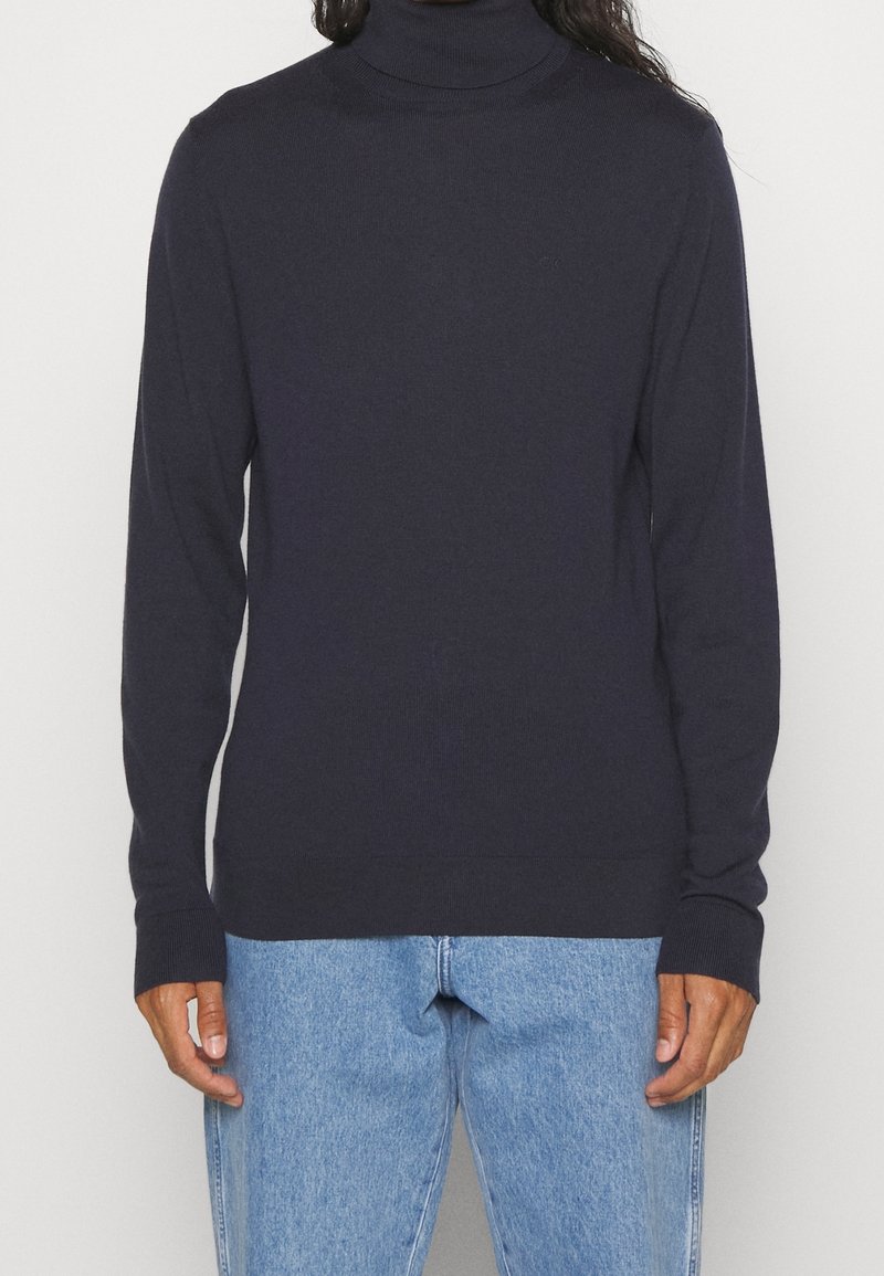Navyfarbener Rollkragenpullover aus weichem, geripptem Material mit langen Ärmeln und körperbetontem Design, kombiniert mit hellblauen Jeans.