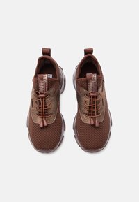 Un paio di sneaker in tessuto a maglia marrone con lacci elastici senza nodi e linguette, viste dall'alto su uno sfondo bianco.