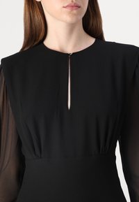 Rochie neagră cu guler înalt, detaliu în formă de cheie la piept și mâneci lungi din material transparent. Țesătură fină, bust ajustat și secțiune la talie plisată.