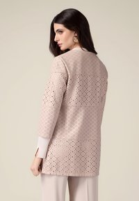 Veste rose clair avec un motif perforé, manches trois-quarts et poignets blancs contrastants. Texture lisse et design structuré.