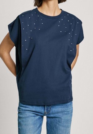 Kobieta w granatowej bluzce bez rękawów ozdobionej srebrnymi koralikami wokół ramion oraz w niebieskich dżinsach.