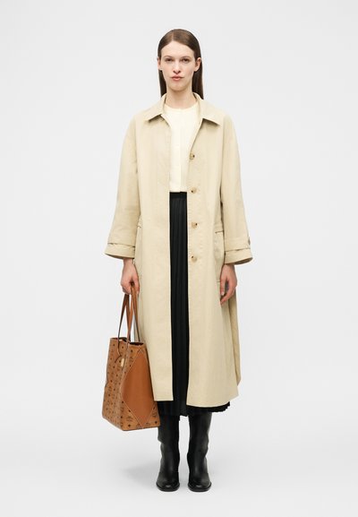 Γυναίκα που φοράει μπεζ μακρύ παλτό, μαύρη πλισέ φούστα, μαύρες μπότες, κρατώντας καφέ τσάντα ώμου με σχέδια, σε λευκό απλό φόντο.