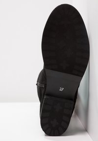 Suela de zapato negra con textura acanalada, tamaño 37 marcado en blanco, contrastando con la parte superior lisa. Diseño minimalista, angulado contra un fondo blanco.