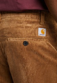 Ciemnobrązowe spodnie z korduroyu z pionowym żeberkowaniem, czarnym zapięciem na guzik oraz białą metką z pomarańczowym logo na tylnej kieszeni.