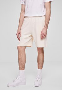 Homme portant un short de sport blanc, des chaussettes blanches montantes, des baskets blanches et un t-shirt blanc à manches courtes, se tenant devant un fond neutre.