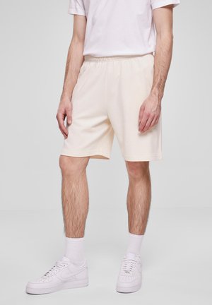 Vyras, vilkintis baltas sportines šortus, baltas trumpas kojines, baltus sportinius batelius ir baltą trumpą rankovę T-shirt, stovi prieš neutralią fono.