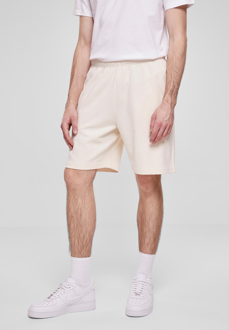 Homme portant un short de sport blanc, des chaussettes blanches montantes, des baskets blanches et un t-shirt blanc à manches courtes, se tenant devant un fond neutre.