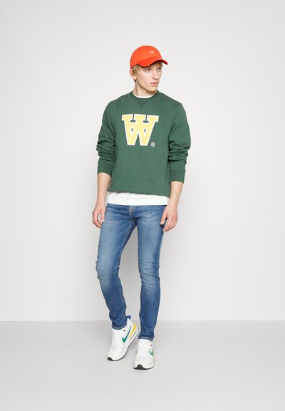 Grüner Sweatshirt mit gelbem "W"-Logo, über einem weißen Shirt getragen. Blaue Skinny-Jeans und weiße Sneaker mit mehrfarbigen Akzenten. Orangene Kappe.
