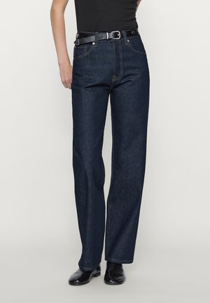 Personne portant un jean droit bleu foncé, une ceinture noire, un haut noir et des chaussures noires, debout devant un fond uni.