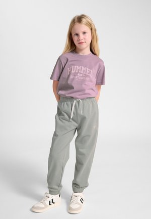 Młoda dziewczyna stoi ze złączonymi rękami z tyłu, ubrana w fioletowy t-shirt, szare spodnie dresowe i białe trampki na jednolitym tle.