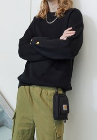 Zwart geribd shirt met lange mouwen met Carhartt-logo, gecombineerd met olijfgroene cargobroeken. Klein zwart met rits afgesloten tasje bevestigd met een clip.