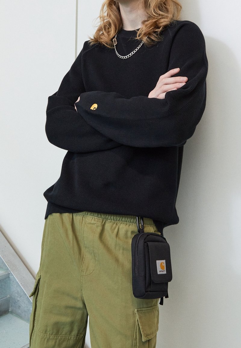 Zwart geribd shirt met lange mouwen met Carhartt-logo, gecombineerd met olijfgroene cargobroeken. Klein zwart met rits afgesloten tasje bevestigd met een clip.