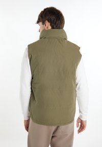 Olivenfarbene gepolsterte Weste mit hohem Kragen, vertikalen Nähten, leichtem Stoff und Seitentaschen, getragen über einem weißen Langarmshirt.