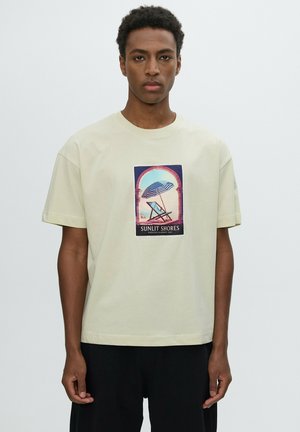 Joven con cabello corto y rizado que lleva una camiseta amarillo pálido con un estampado de una silla de playa y una sombrilla con el texto "Sunlit Shores".