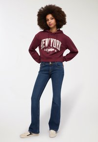Bordowy, skrócony hoodie z nadrukiem "NEW YORK", w połączeniu z rozszerzanymi, ciemnoniebieskimi dżinsami i kremowymi trampkami. Swobodny, luźny krój.