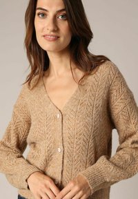 Beige gebreide cardigan met een V-hals, voorzien van een gestructureerd ruitpatroon, lange mouwen en witte knopen aan de voorkant.